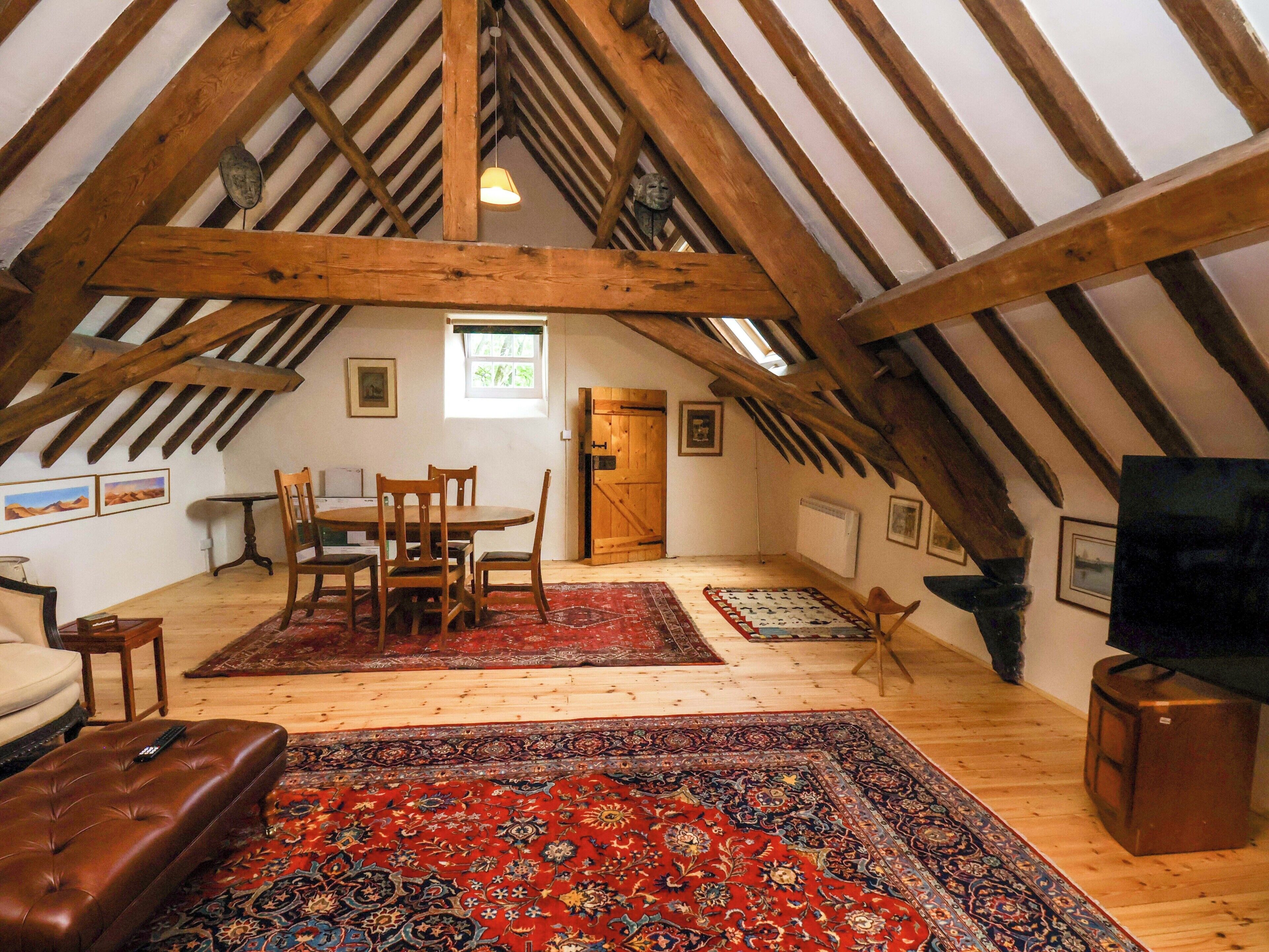 Een loft-achtig vertrek met een hoge plafond, houten balken, een eettafel met stoelen, een bank, een televisie en een kamerbreed tapijt met dessin.