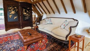 A room with a high ceiling, exposed wooden beams, a sofa, a cushioned ottoman, a traditional rug, and a dark wooden cabinet with a painting. - The Gardener's Cottage, Low Hall (Scarborough)