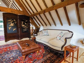 A room with a high ceiling, exposed wooden beams, a sofa, a cushioned ottoman, a traditional rug, and a dark wooden cabinet with a painting. - The Gardener's Cottage, Low Hall (Scarborough)