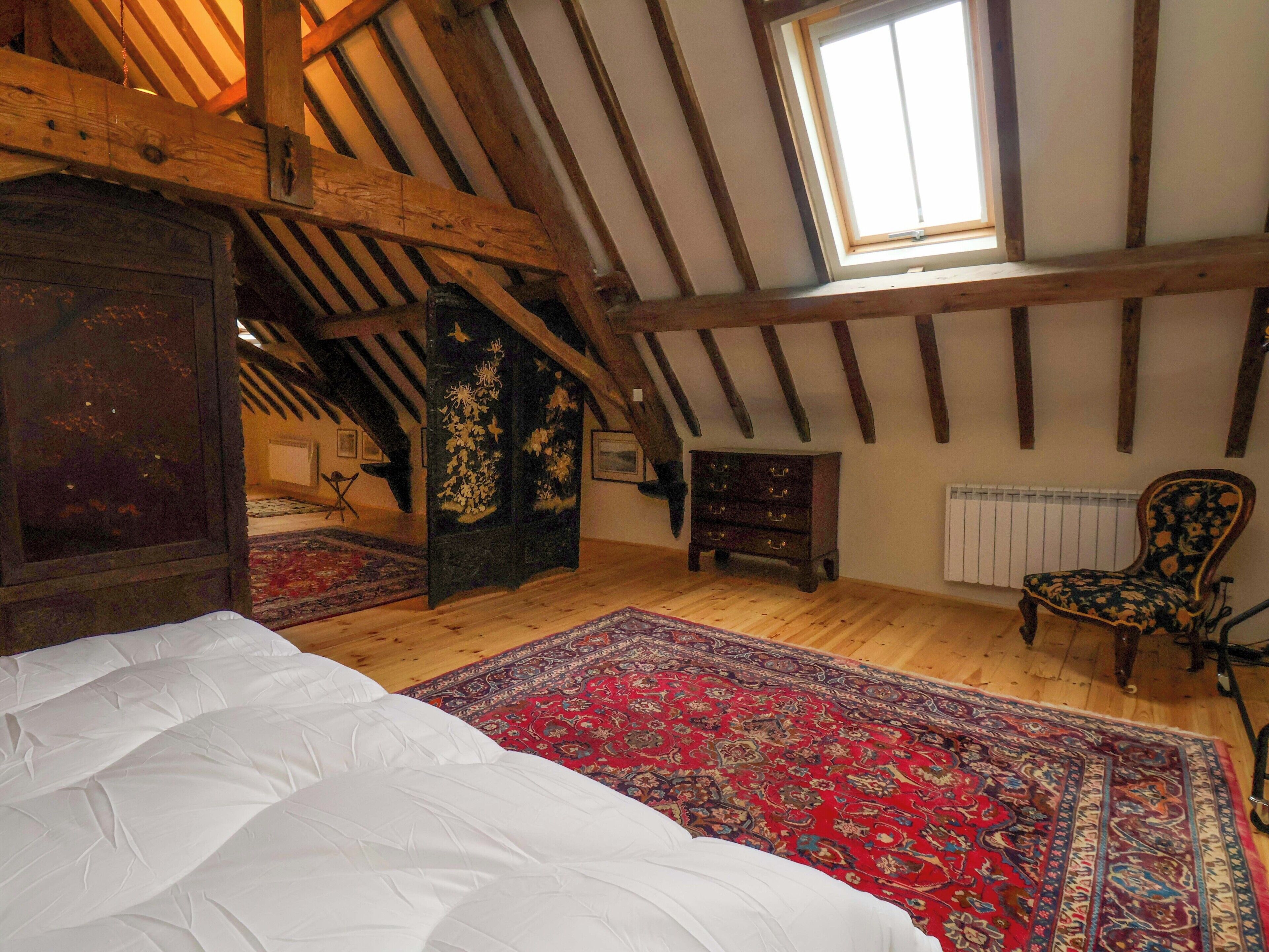Una habitación con techos altos y vigas de madera a la vista, una ventana grande, una cama, un tocador, una silla y una alfombra estampada.