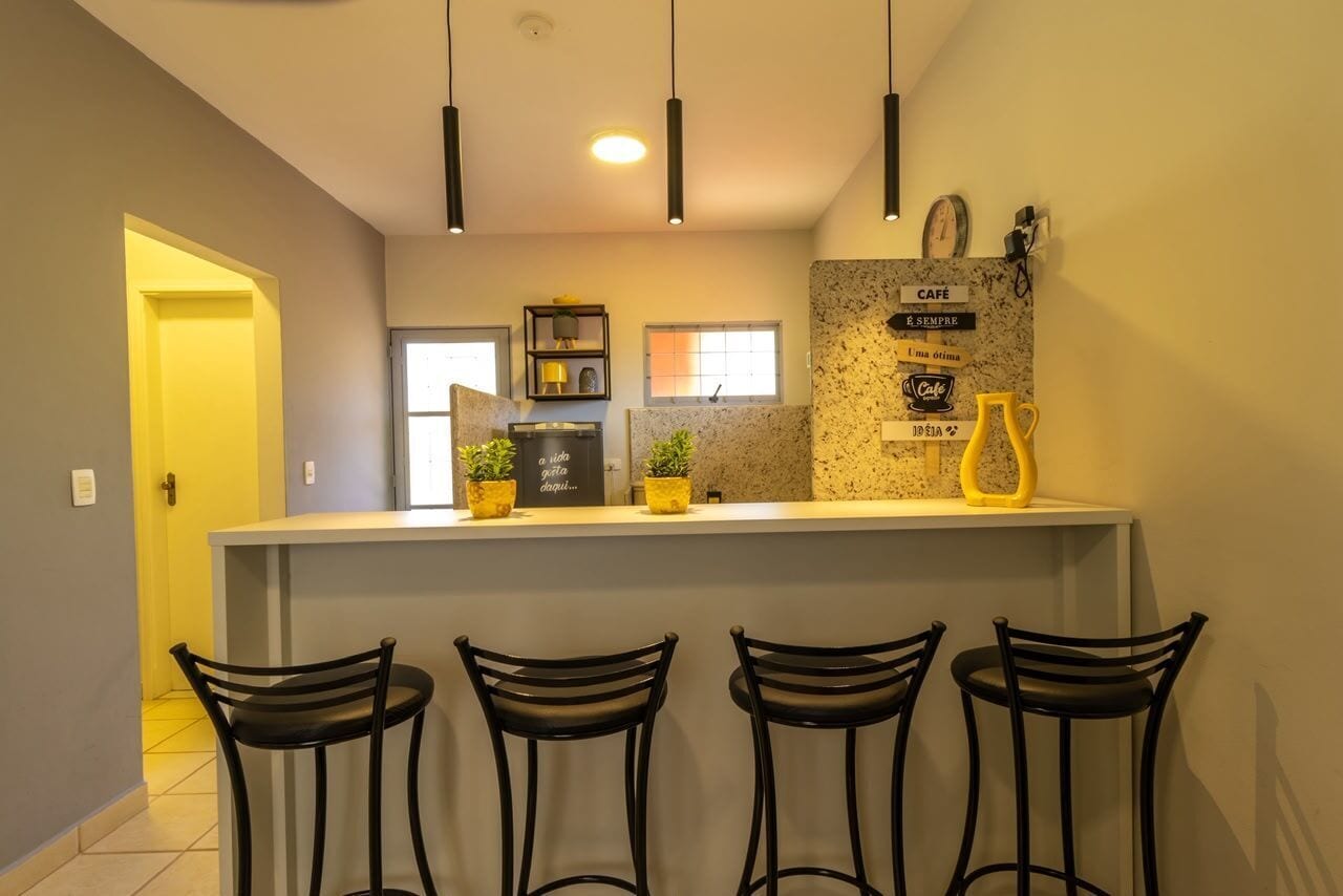 Un área de cocina con barra, tres taburetes negros, luces colgantes, un reloj y un cartel que dice 'CAFE'.