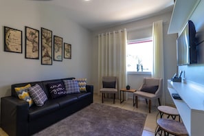 Un salon moderne avec un canapé en cuir noir, deux fauteuils, une table basse, un téléviseur à écran plat et une grande fenêtre avec des rideaux.