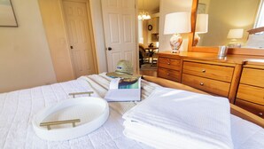 Una cama bien hecha con edredón blanco, una bandeja redonda blanca y una toalla blanca doblada. Hay un aparador de madera con espejo y lámpara. Se ve una puerta al fondo que conduce a otra habitación.