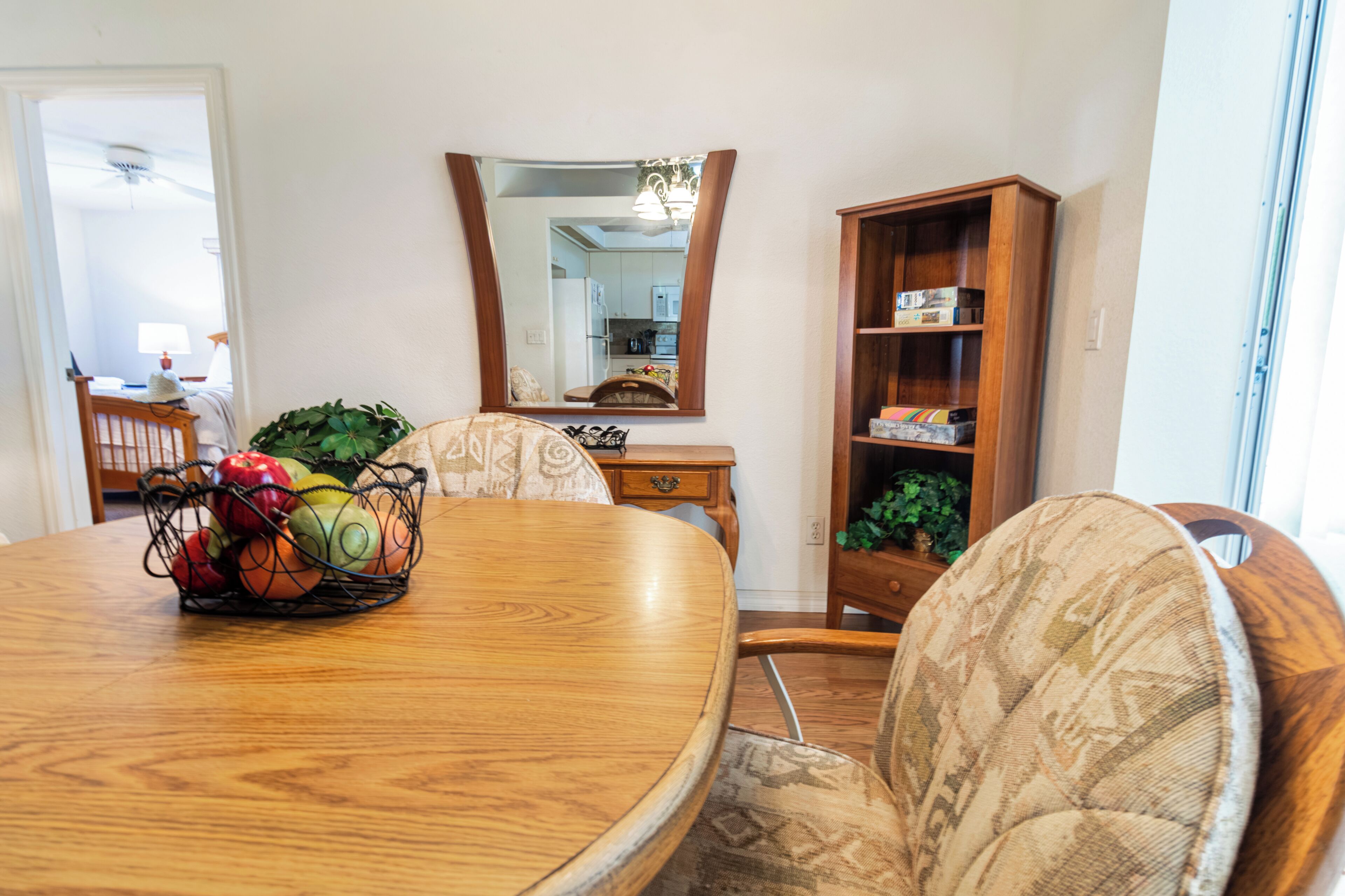 Un comedor con una mesa de madera, sillas, una cesta de frutas, un espejo y una estantería.