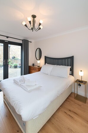 Ein ordentlich bezogenes Bett mit weißen Leinen, eine Nachttischlampe, ein Holfnachtkästchen und ein großer Spiegel an der Wand.