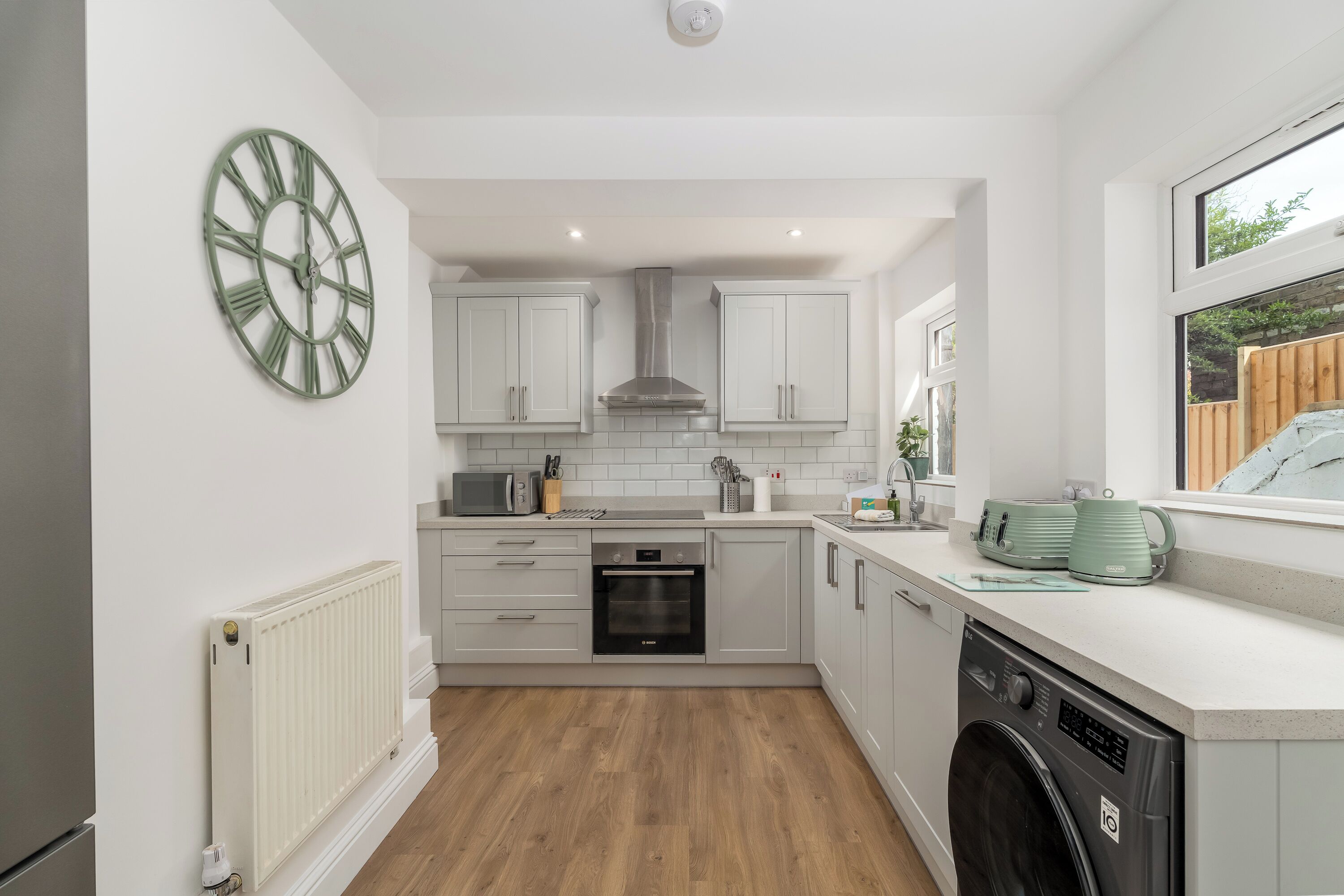 A modern kitchen with white cabinets, a built-in oven, a washing machine, and a large green clock on the wall.