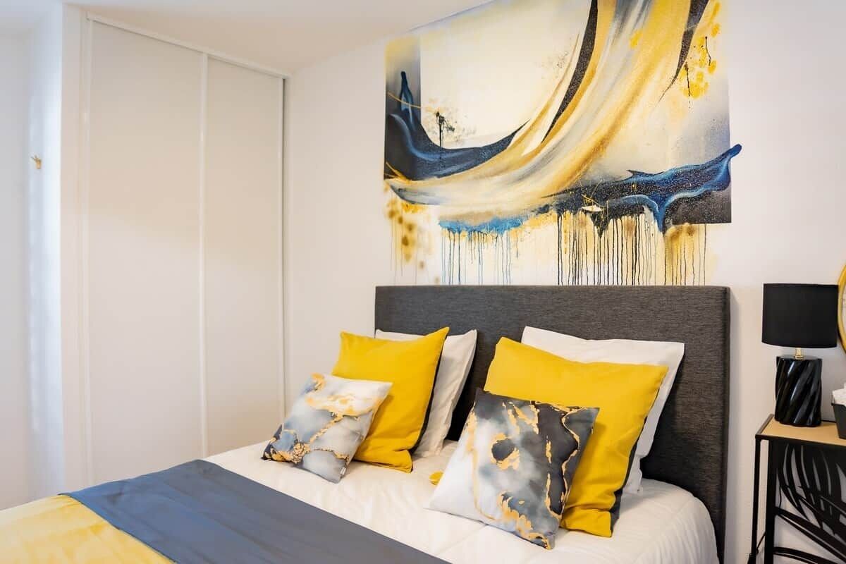 Een slaapkamer met een bed, kussens, een hoofdbord, een schilderij en een nachtkastje.