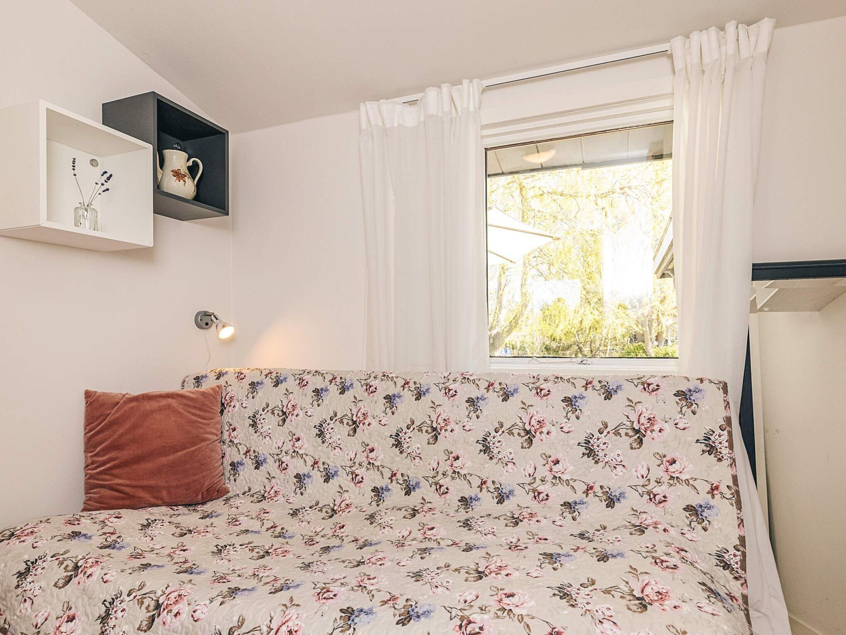 A bed with floral bedding, a brown pillow, a bedside table with a lamp, and a window with curtains.