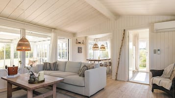 A spacious room with a light-colored sofa, a wooden coffee table, wicker chairs, and a dining table with hanging lights.