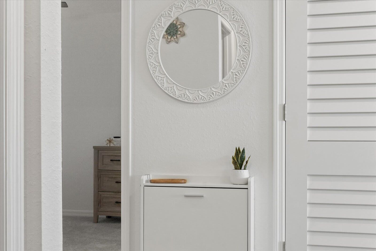 Un pasillo con una puerta blanca, un aparador blanco con una planta y un espejo decorativo en la pared.