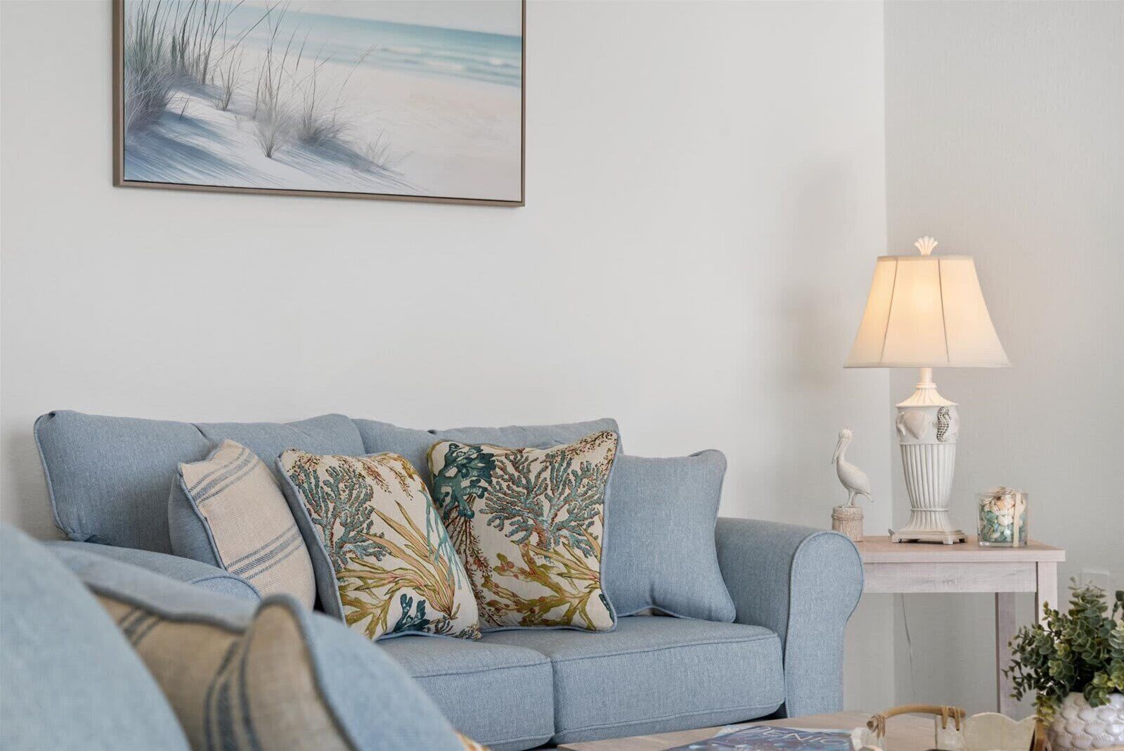 Una sala de estar con un sofá azul claro, cojines estampados, una obra de arte enmarco con una playa, una lámpara blanca y una pequeña mesita con objetos decorativos.
