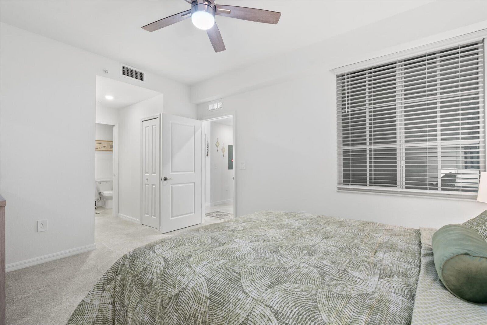 A bedroom with a ceiling fan, a bed with a patterned bedspread, a window with blinds, and a door leading to another room.