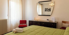 Quarto de hotel com cama, uma cadeira vermelha, cômoda com espelho, lustre e janela com cortinas.
