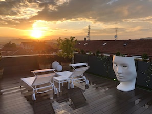 A rooftop terrace with white lounge chairs, a white bust sculpture, and a view of the sunset over a cityscape. - Exclusive penthouse in the historic center of Alba, panoramic terrace with garden and private  pool and solarium. (alba)