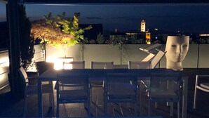 An outdoor dining area with chairs and tables, a pool with blue lighting, and a view of a cityscape with a tower in the background. - Exclusive penthouse in the historic center of Alba, panoramic terrace with garden and private  pool and solarium. (alba)