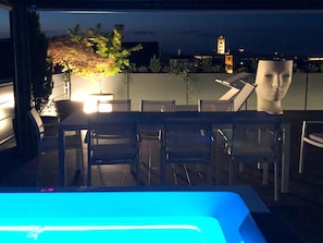 An outdoor dining area with chairs and tables, a pool with blue lighting, and a view of a cityscape with a tower in the background. - Exclusive penthouse in the historic center of Alba, panoramic terrace with garden and private  pool and solarium. (alba)