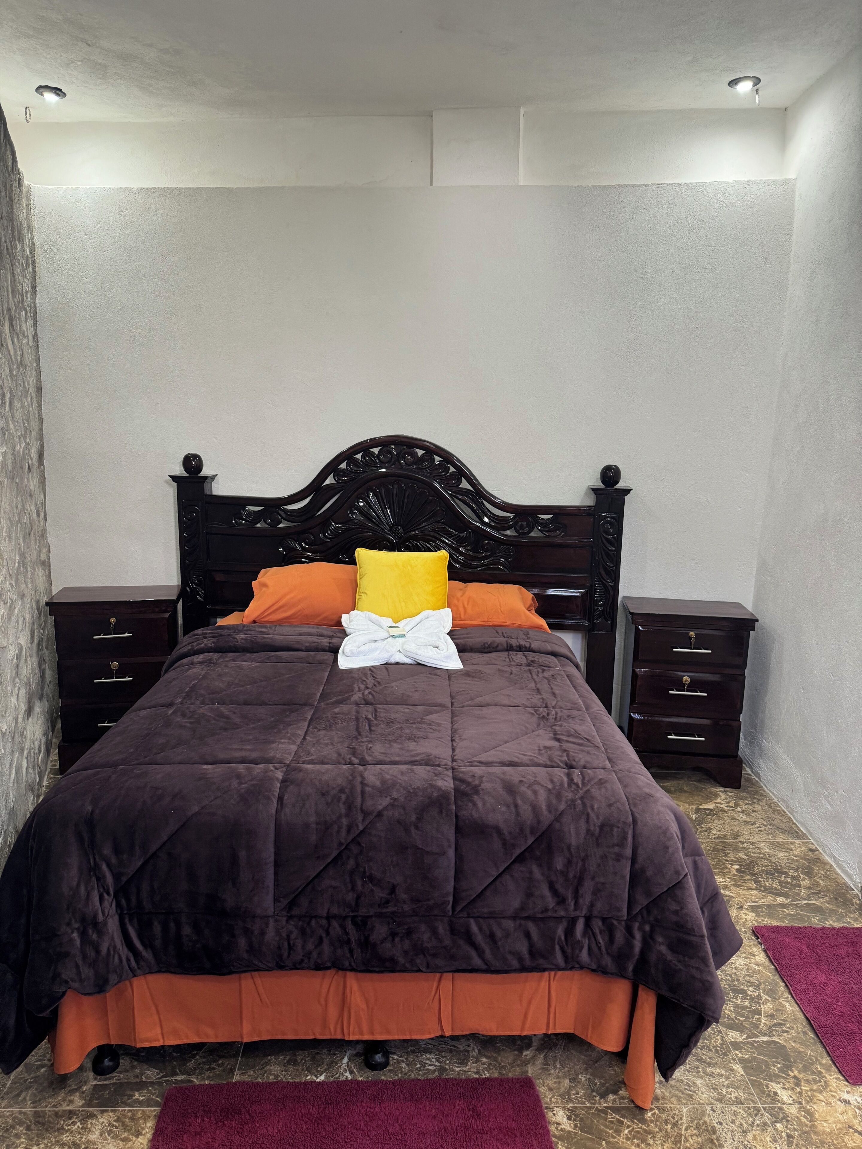 Ein ordentlich bezogenes Bett mit dunklem Kopfteil, einer braun-orangenen Tagesdecke und einem weißen Handtuch. Zwei Nachttische aus Holz mit Schubladen, ein violetter Teppich auf dem Boden und eine strukturierte Wand sind ebenfalls zu sehen.