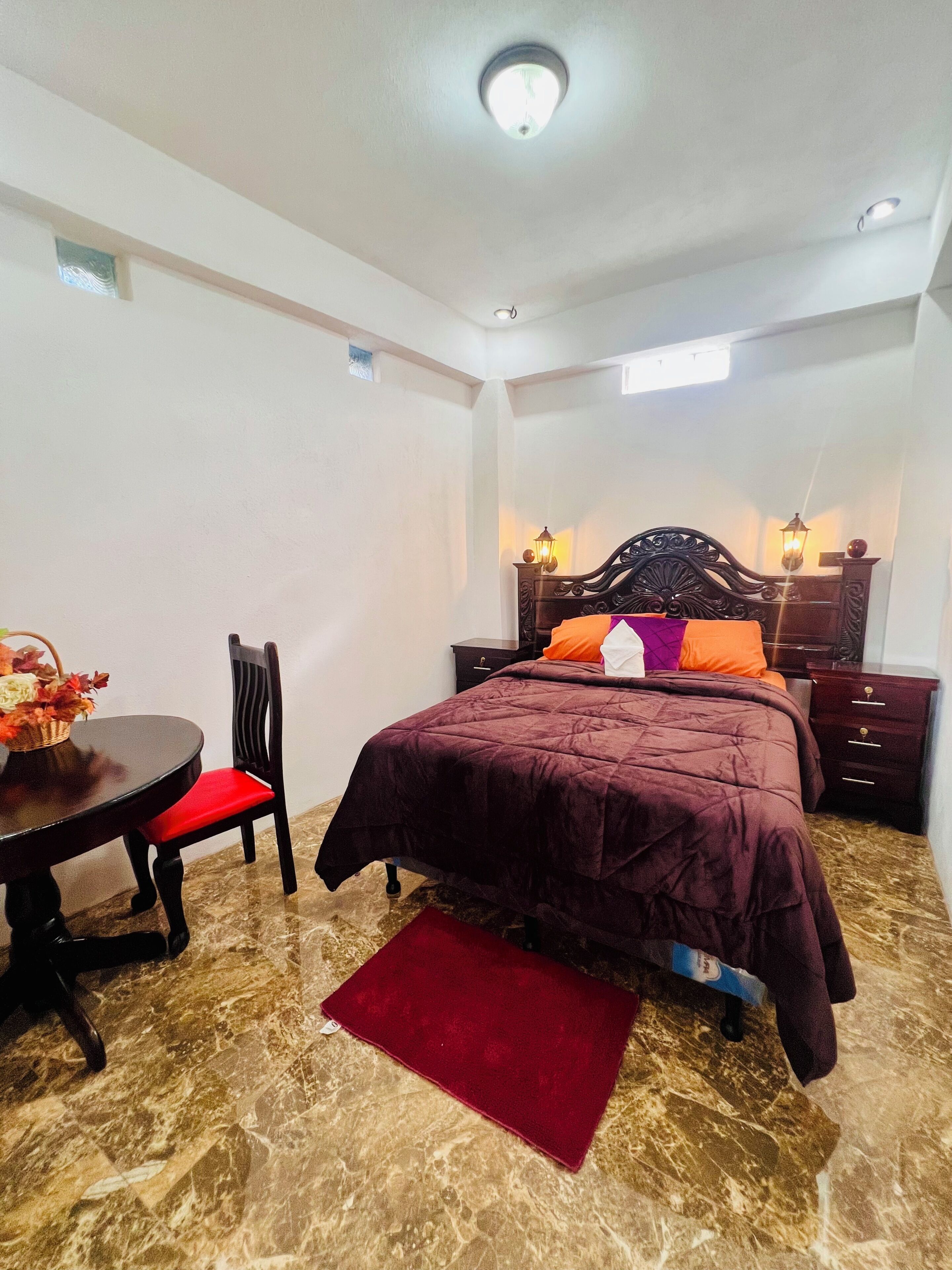 Un dormitorio con cama, una mesa con silla, un tapete rojo y una mesita de noche con lámpara.