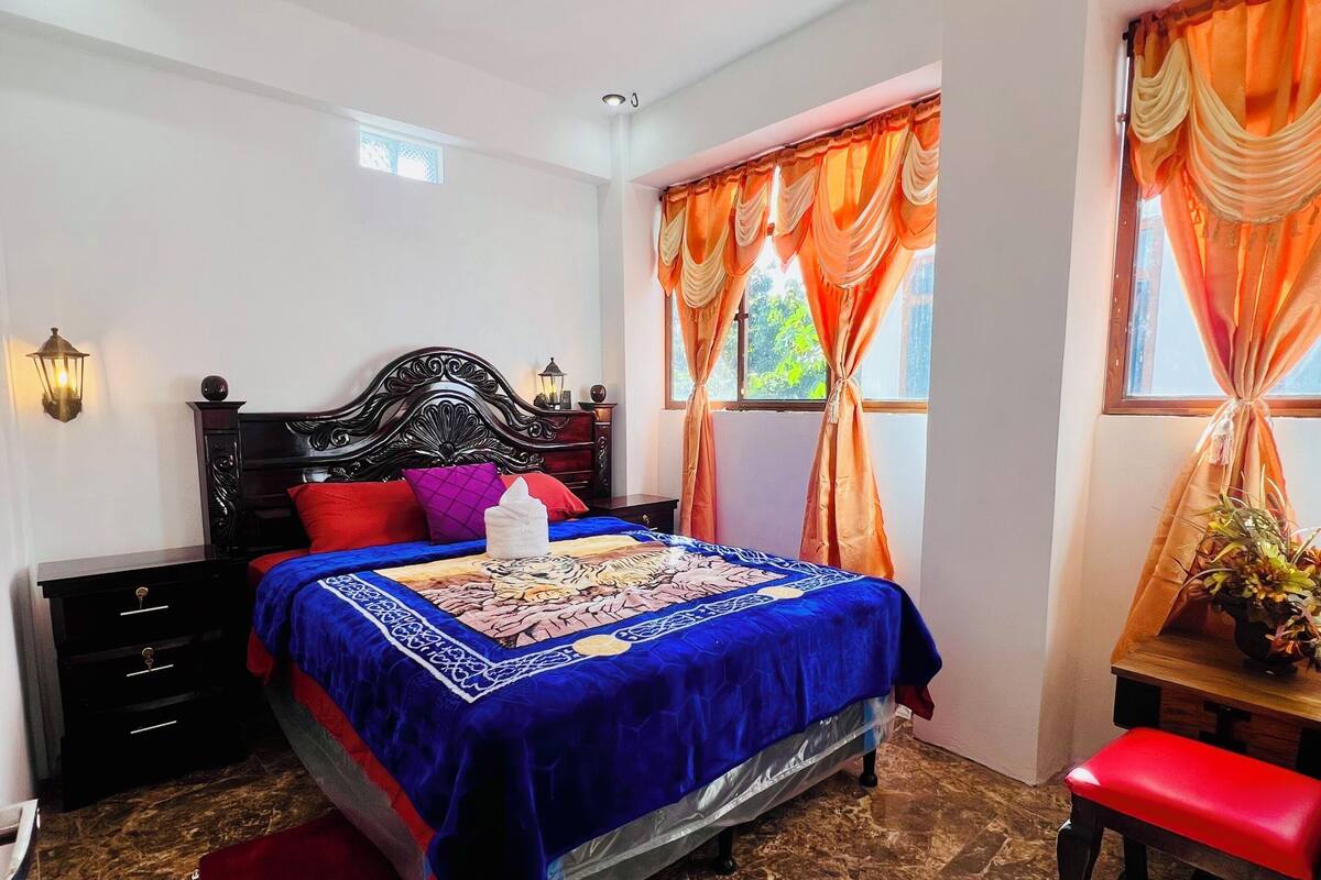 A bedroom with a bed, bedside tables, a nightstand, a bench, a window with curtains, and a ceiling light.