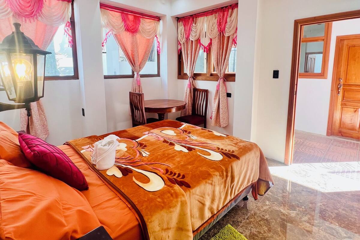 A bedroom with a bed, pillows, a nightstand, a lamp, and a window with curtains.