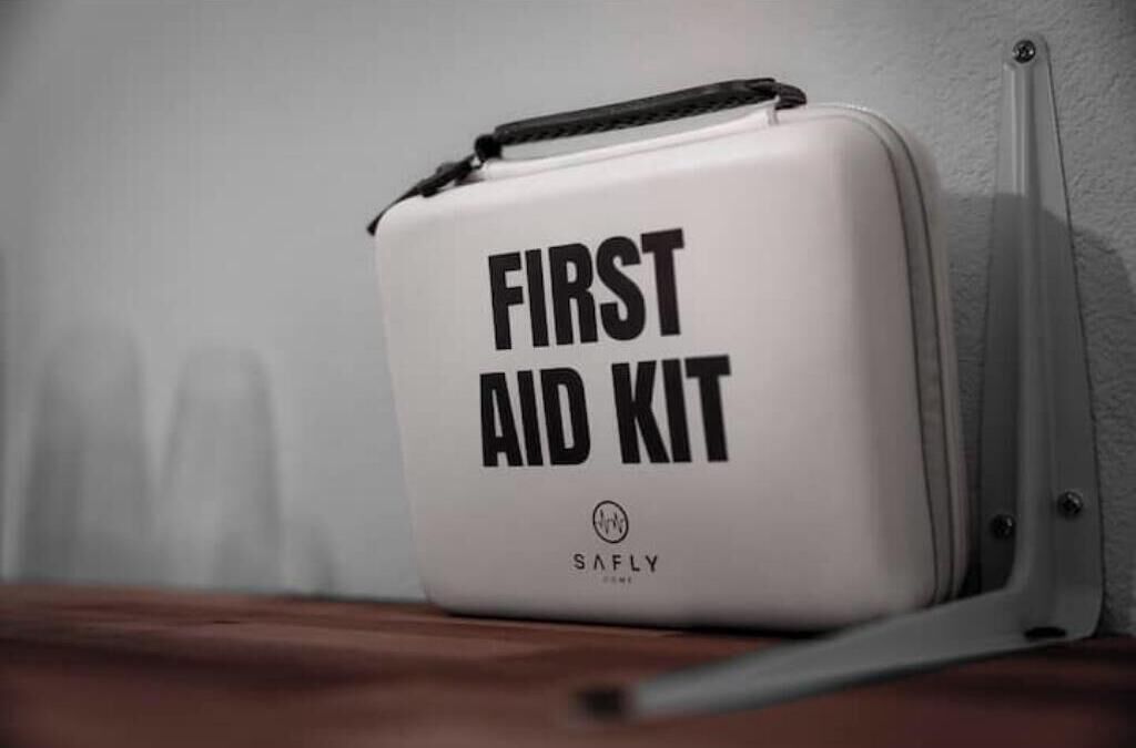 A white case labeled 'FIRST AID KIT' with a logo, placed on a wooden surface against a wall.