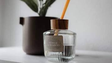 Una botella de vidrio transparente con un palito de madera, sobre una superficie blanca, con una planta en maceta al fondo.