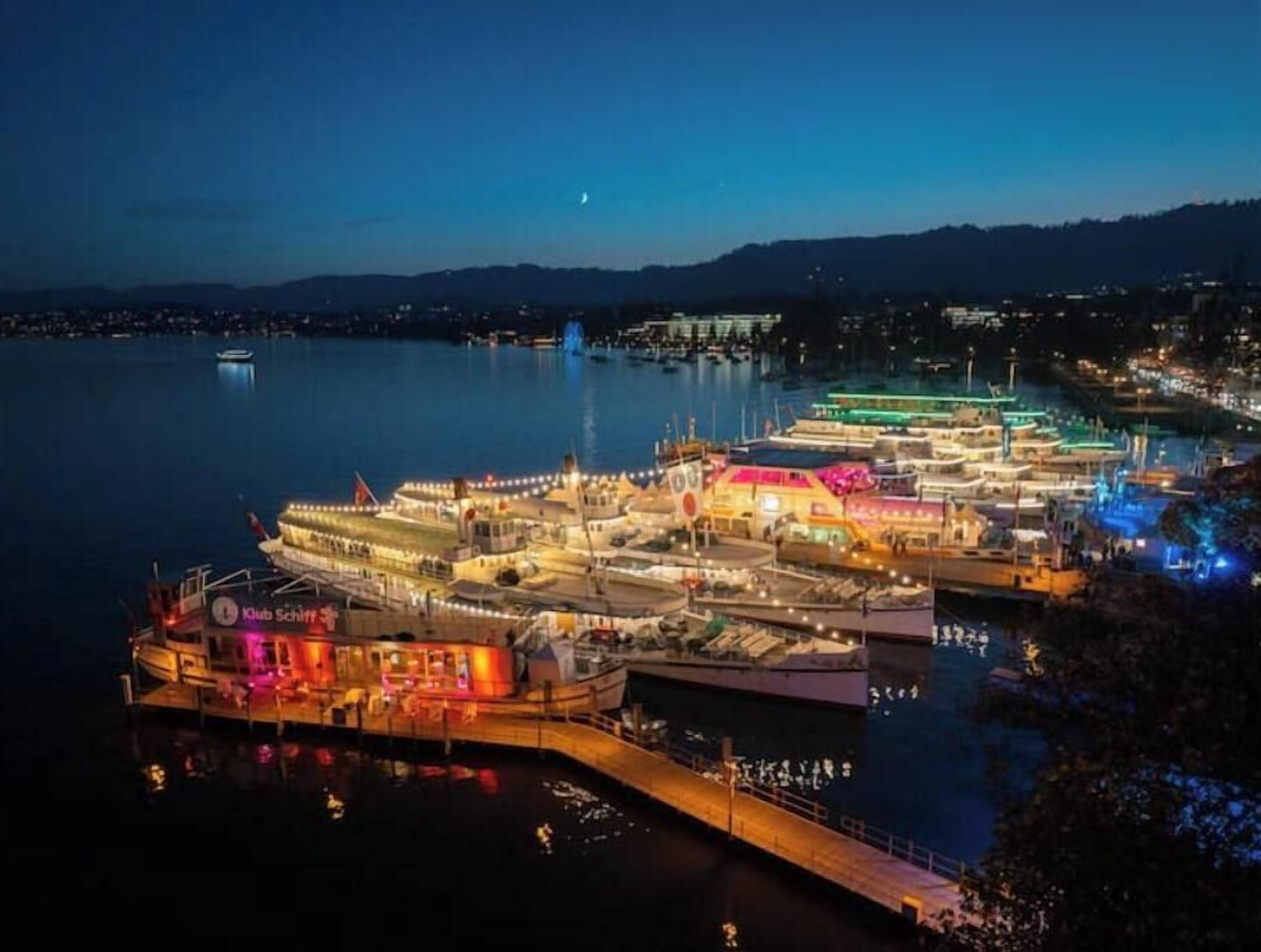 A marina with multiple boats docked, a pier extending into the water, and a cityscape in the background.