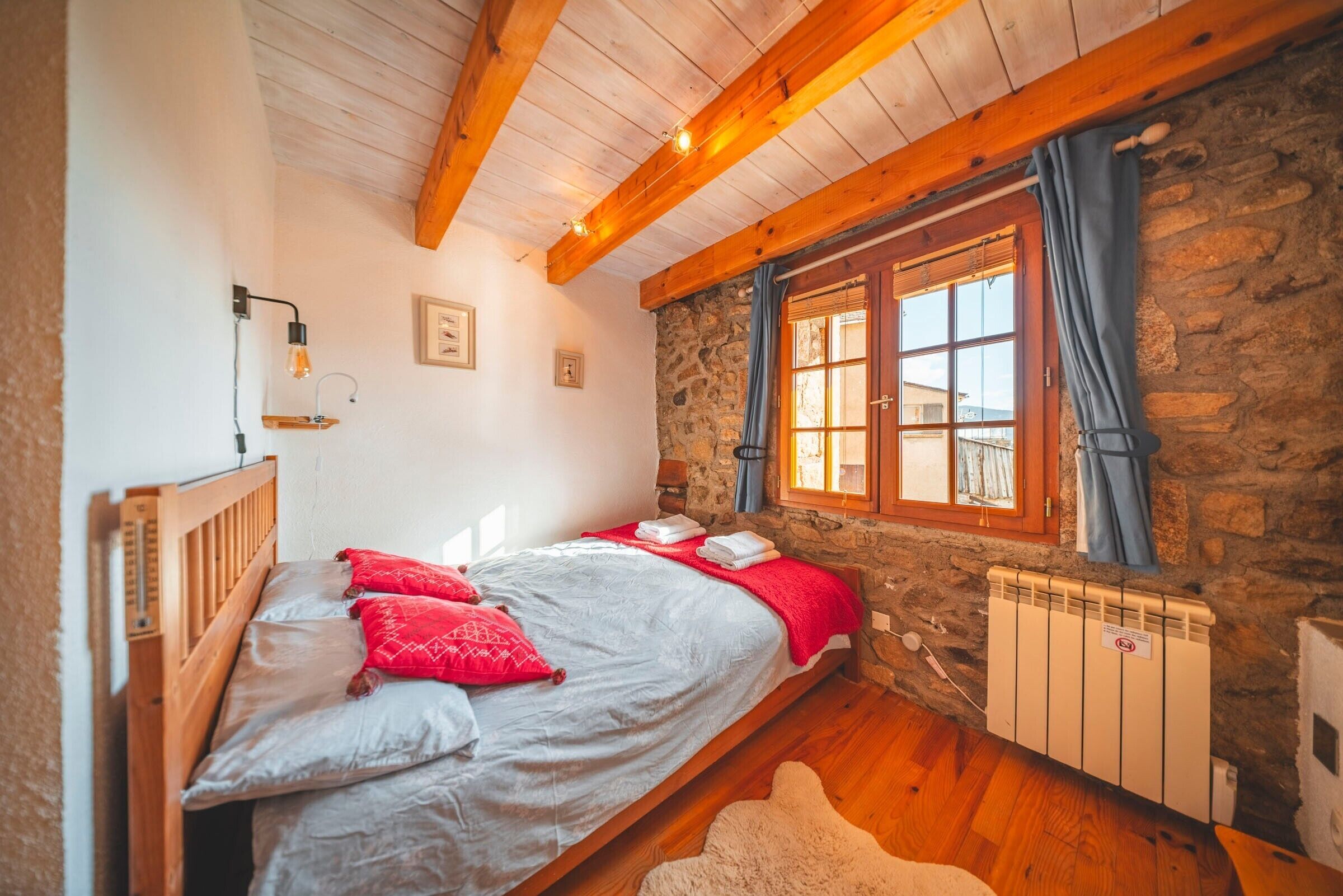 A bedroom with a stone wall, wooden ceiling beams, a bed with red and white bedding, a window with curtains, and a radiator.