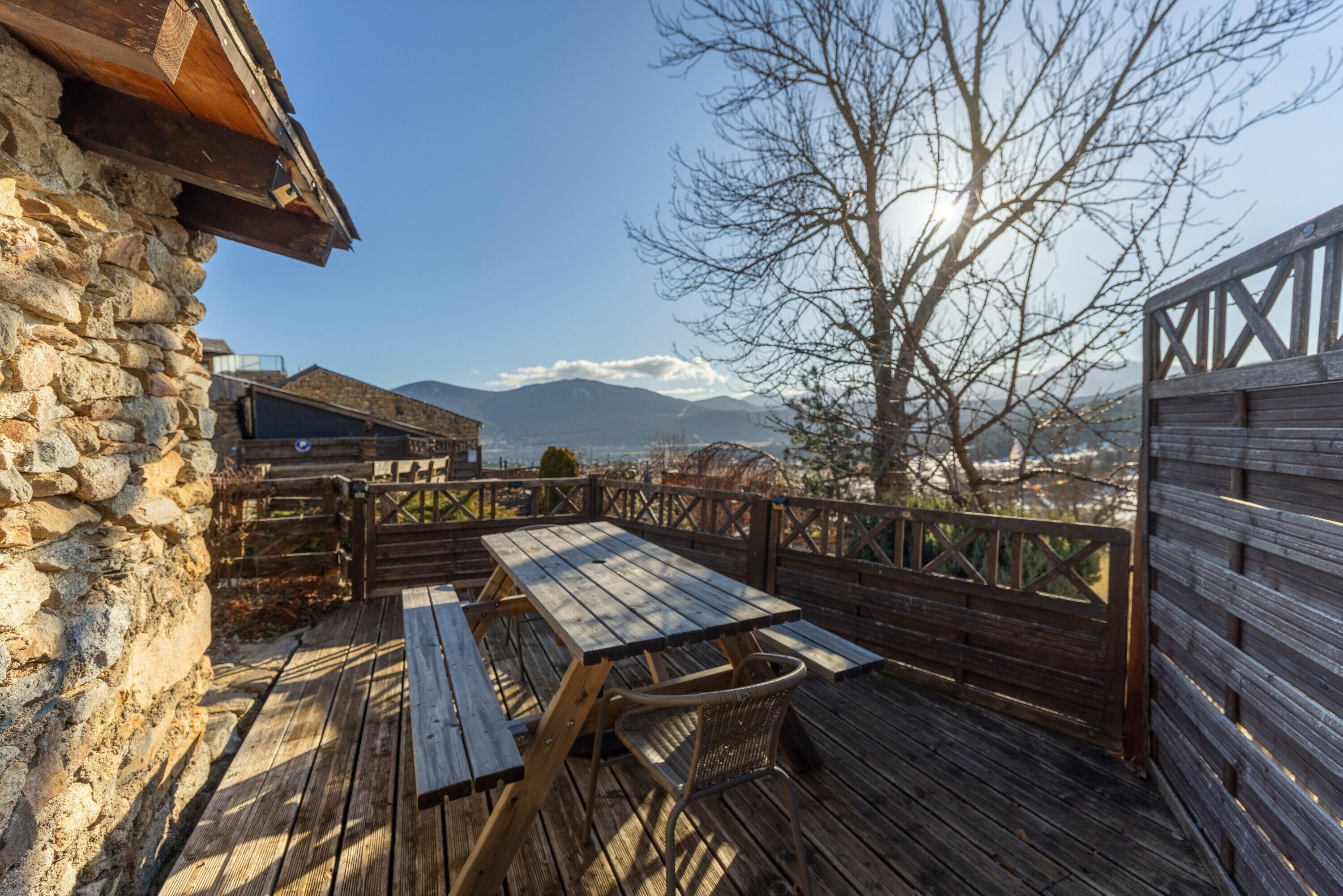 Terraza de madera con mesa y sillas, paredes de piedra y vista a las montañas.