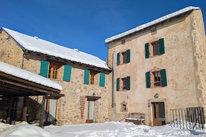 A stone building with snow-covered roofs and green shutters, a wooden door, and a small balcony. - La Grange de la Petite Ourse with Large Furnished Terrace, Stunning Lake and Mountain Views (Les Angles)