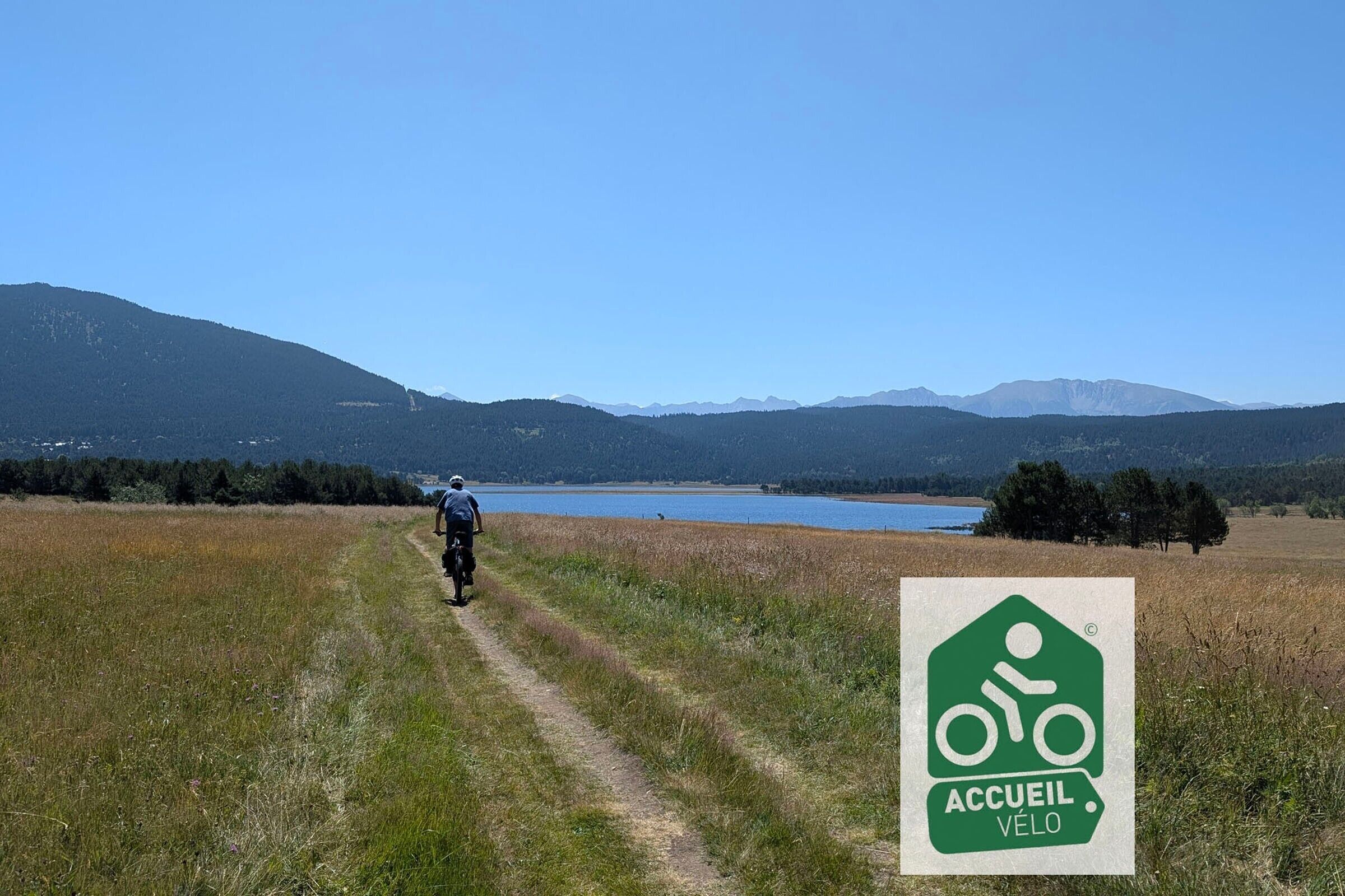 Een pad dat naar een meer leidt, met bergen op de achtergrond en een groen fietssymbool dat aangeeft dat het gebied geschikt is voor fietsers.