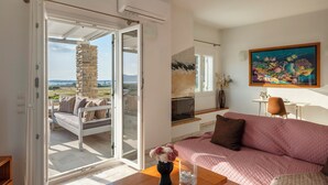 A living room with a sofa, a table with a vase, a painting, and a view through a glass door. - Where the sea meets the sky your dream home in Paros with a view that never ends (Kampos)