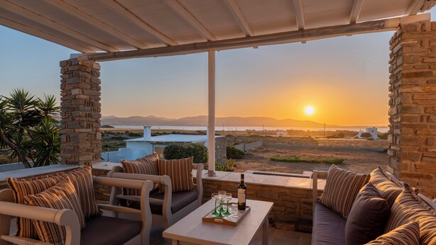 A terrace with outdoor seating, a table with glasses and a bottle, and a view of the sunset over a landscape. - Where the sea meets the sky your dream home in Paros with a view that never ends (Kampos)