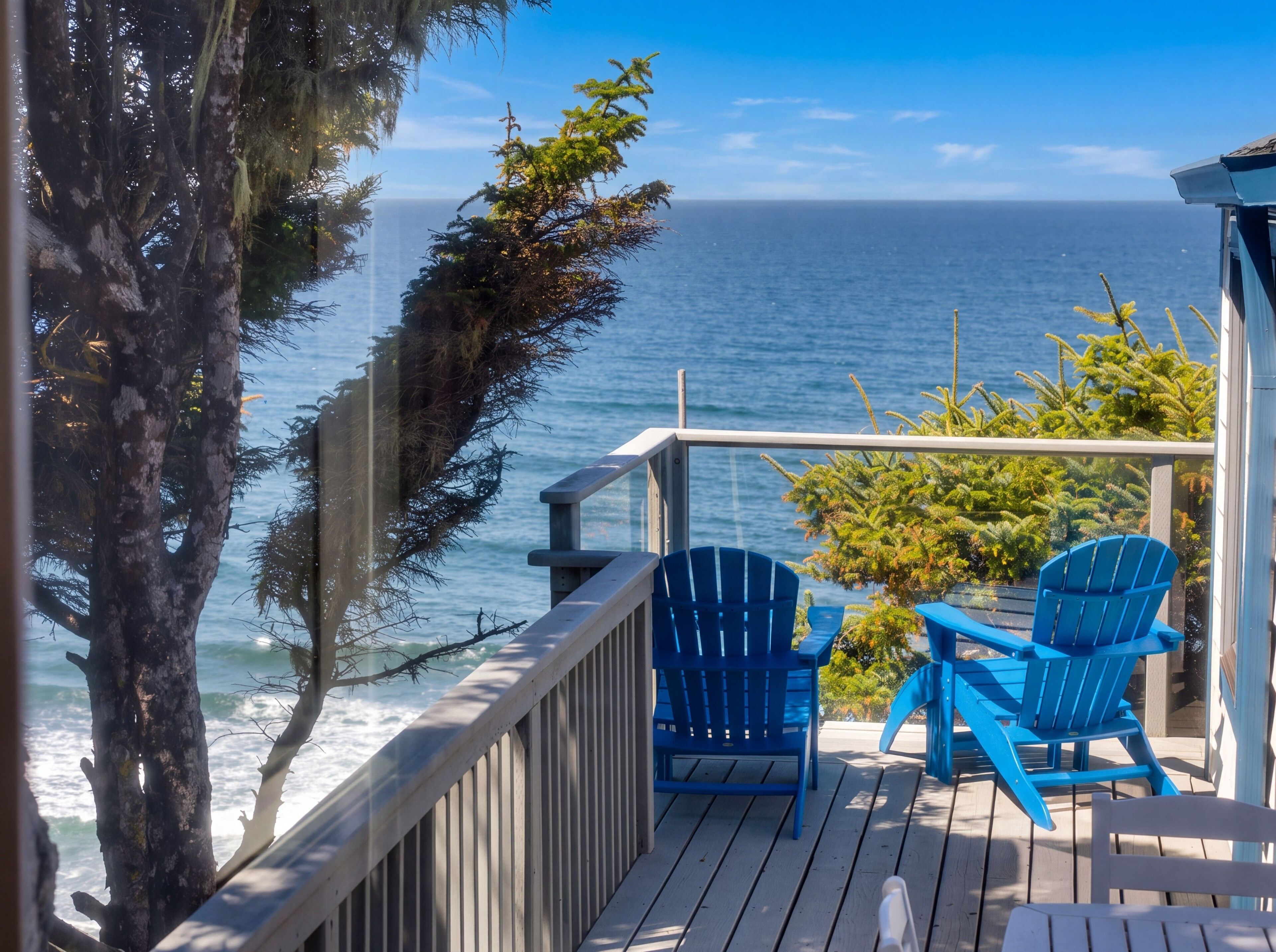Una terraza de madera con dos sillas azules y una mesa, con vista a una playa y olas del océano bajo un cielo despejado.