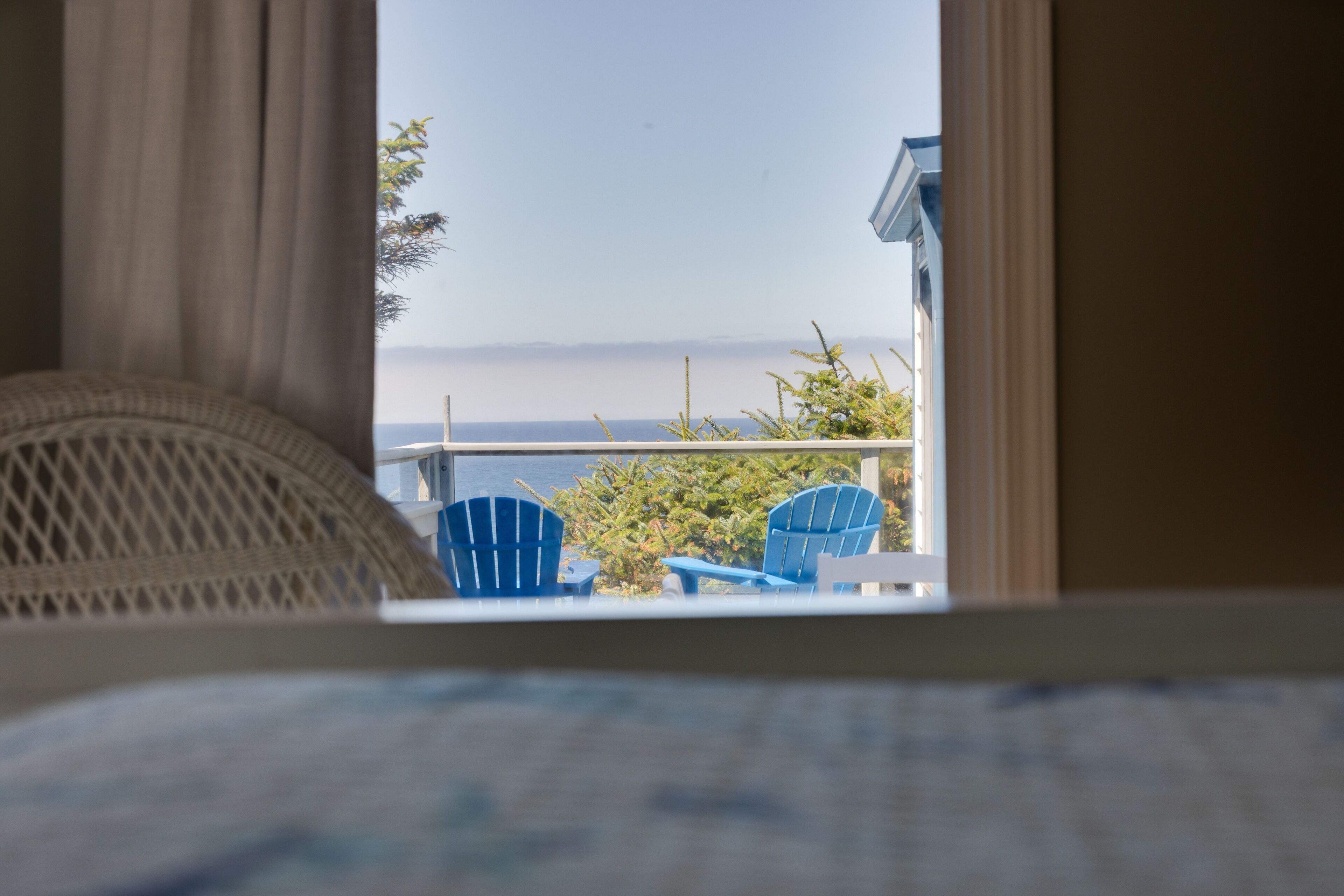 Uitzicht vanuit een raam, met een rieten stoel en twee blauwe stoelen buiten, op een wateroppervlak en een verre kustlijn.