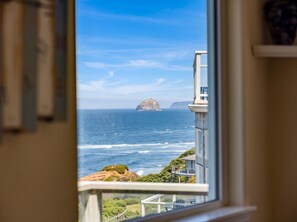 A coastal view from a window overlooking the sea, a rocky island, and a building with a balcony. - Pelican's Perch at Neskowin Ocean View (Cloverdale)