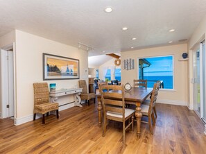 A dining area with a wooden table and chairs, a framed picture on the wall, and a view of the ocean through a large window. - Pelican's Perch at Neskowin Ocean View (Cloverdale)