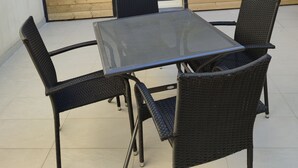 En udendørs terrasse med et bord, stole, en trævæg og et vindue.