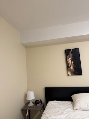A bedroom with a bed, a bedside table with a lamp and a digital clock, and a wall-mounted painting.
