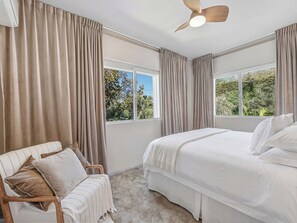 Ein Schlafzimmer mit Bett, Stuhl, Deckenventilator und großen Fenstern mit Vorhängen.