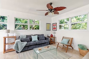 Ein Wohnzimmer mit einer Couch, einem Glastisch, einem Holzstuhl, einem Deckenventilator und großen Fenstern mit Blick auf Grünflächen.