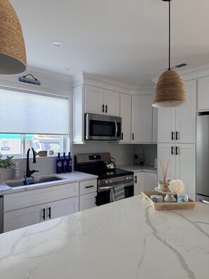 A modern kitchen with white cabinetry, stainless steel appliances, a marble countertop, and a hanging woven light fixture. - Beautiful 3 Bedroom Cottage steps from the Beach with brand new Trex deck (East Lyme)