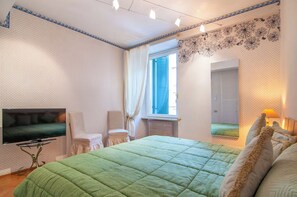 A bedroom with a bed, a TV, a chair, a window with curtains, and a wall with a floral pattern. - [Desiderium Home] A stone's throw from the Arena, Juliet's House, Roman Theater (Verona)