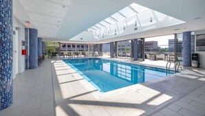 A modern indoor swimming pool with a skylight ceiling, clear views of the city, and a tiled floor. - Downtown Stamford Luxury | Indoor Pool & Gym (Stamford, CT)