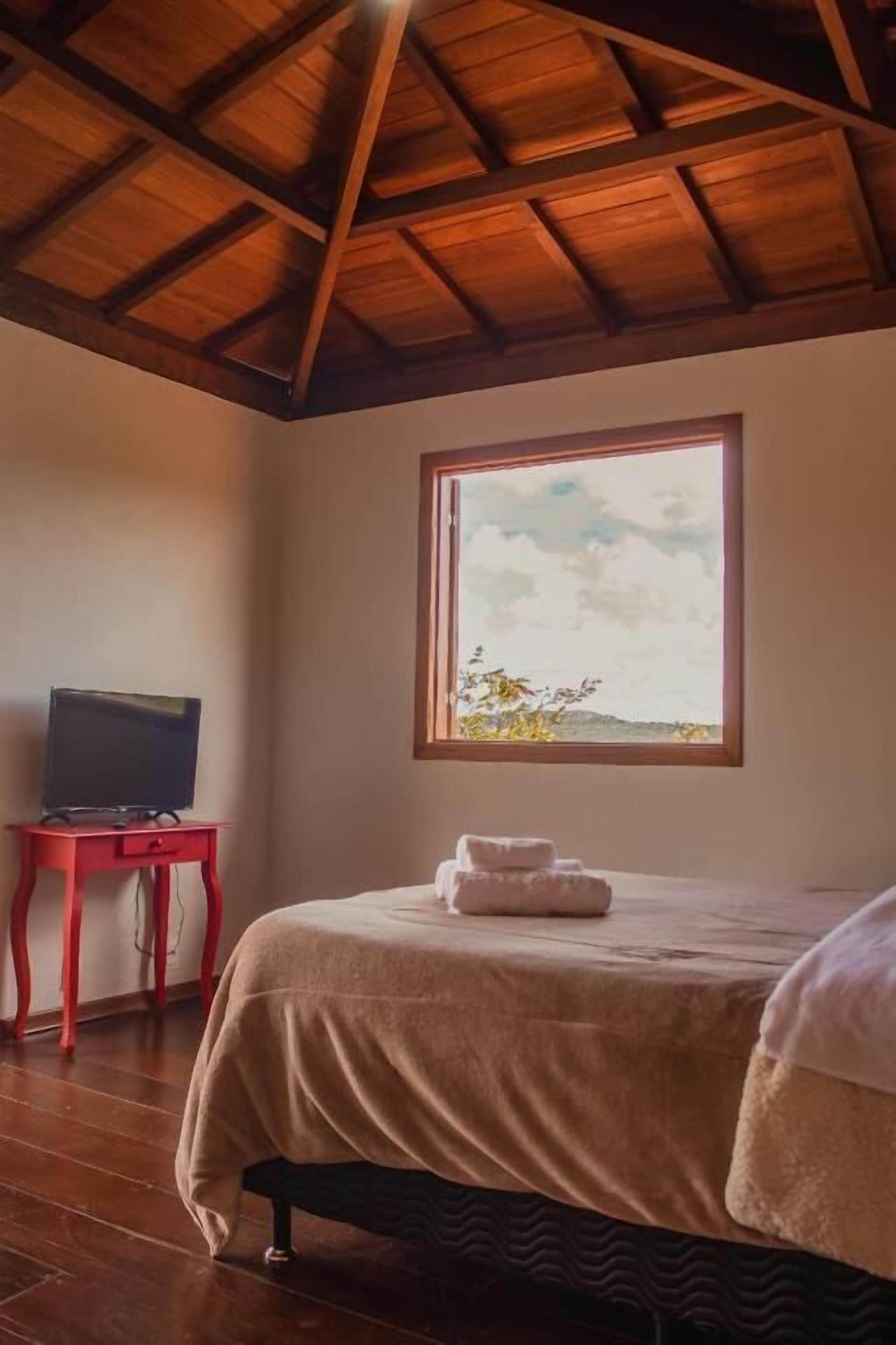 Ein Zimmer mit Holzboden, einem Bett mit weißer Bettdecke, einem roten Tisch mit Fernseher und einem Fenster mit Blick ins Freie.