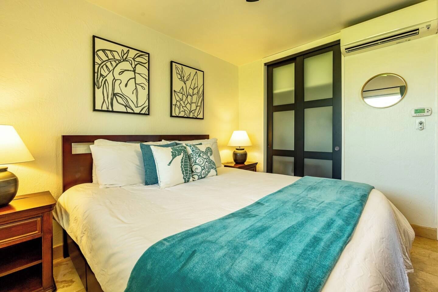 A hotel room with a bed, two wall-mounted artworks, a nightstand with a lamp, and a door with a frosted glass panel.
