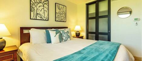 A hotel room with a bed, two wall-mounted artworks, a nightstand with a lamp, and a door with a frosted glass panel.