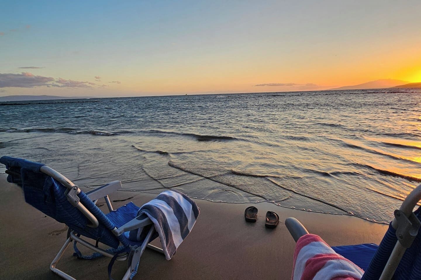 A beach scene with a chair and a pair of shoes on the sand, facing the ocean at sunset.