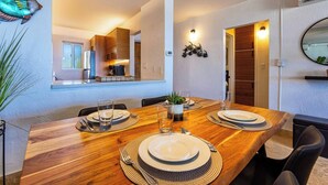Comedor con mesa de madera dispuesta para cuatro, cocina al fondo y un espejo colgado en la pared.