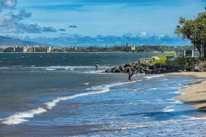 Ein Strand mit Wellen, felsiger Küste und Gebäuden im Hintergrund.